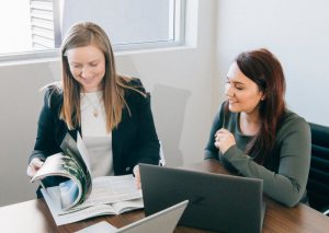 Two women discussing while checking a magazine and working in laptop